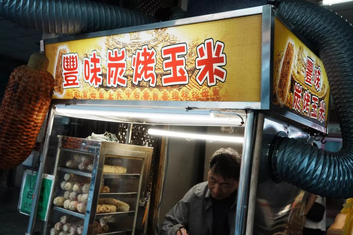 【板橋裕民夜市】《豐味碳烤玉米》評價:一半是炭火靈魂的醬香,另一半是米酒香氣的人情味 10 豐味碳烤玉米的攤位正面,黃色招牌上有店名,老闆正在攤位內專注地備料,旁邊的玻璃櫃中放著待烤的玉米。