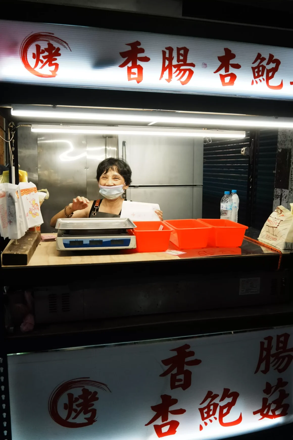 【板橋裕民夜市】《豐味碳烤玉米》評價:一半是炭火靈魂的醬香,另一半是米酒香氣的人情味 13 豐味碳烤玉米的老闆娘戴著口罩在攤位裡工作,她從櫃檯後方抬頭親切地看著鏡頭,身旁有著寫著「杏鮑菇」的招牌。