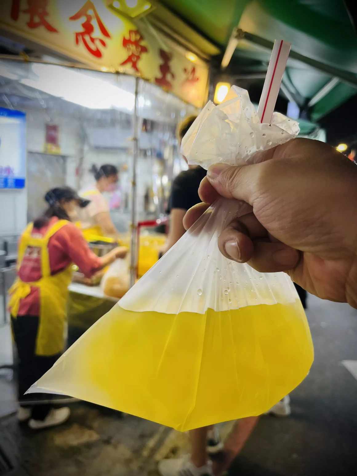 【萬華夜市】《懷念愛玉冰》:一半是回憶的召喚術,另一半是長大後的誠實豆沙包 17 一隻手在萬華夜市裡,拿著一袋用傳統塑膠袋裝的懷念愛玉冰,袋中插著吸管,背景是模糊的店家攤位,充滿懷舊氛圍。