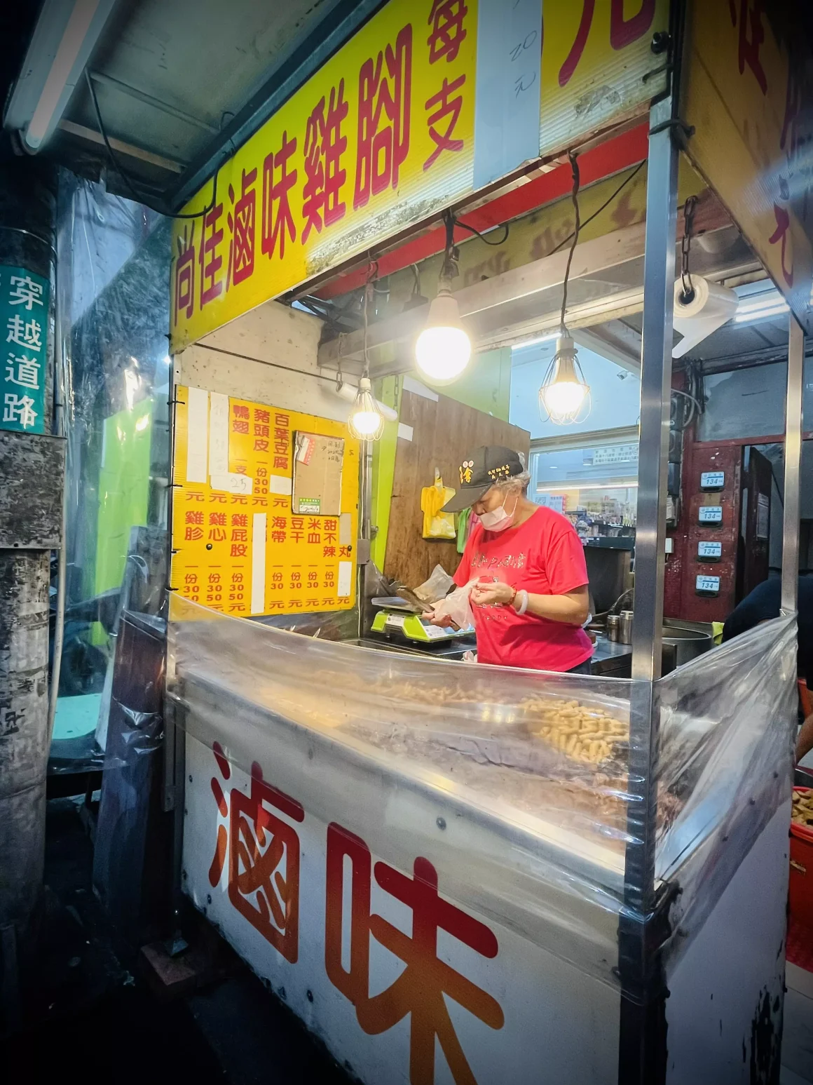 【板橋】《尚佳滷味》評價:一半是錢包的摯友,另一半是追劇的稱職配角 8 板橋尚佳滷味攤的夜間全景,老闆正戴著口罩在燈火通明的攤位內為客人準備滷味,黃色的招牌與價目表清晰可見。