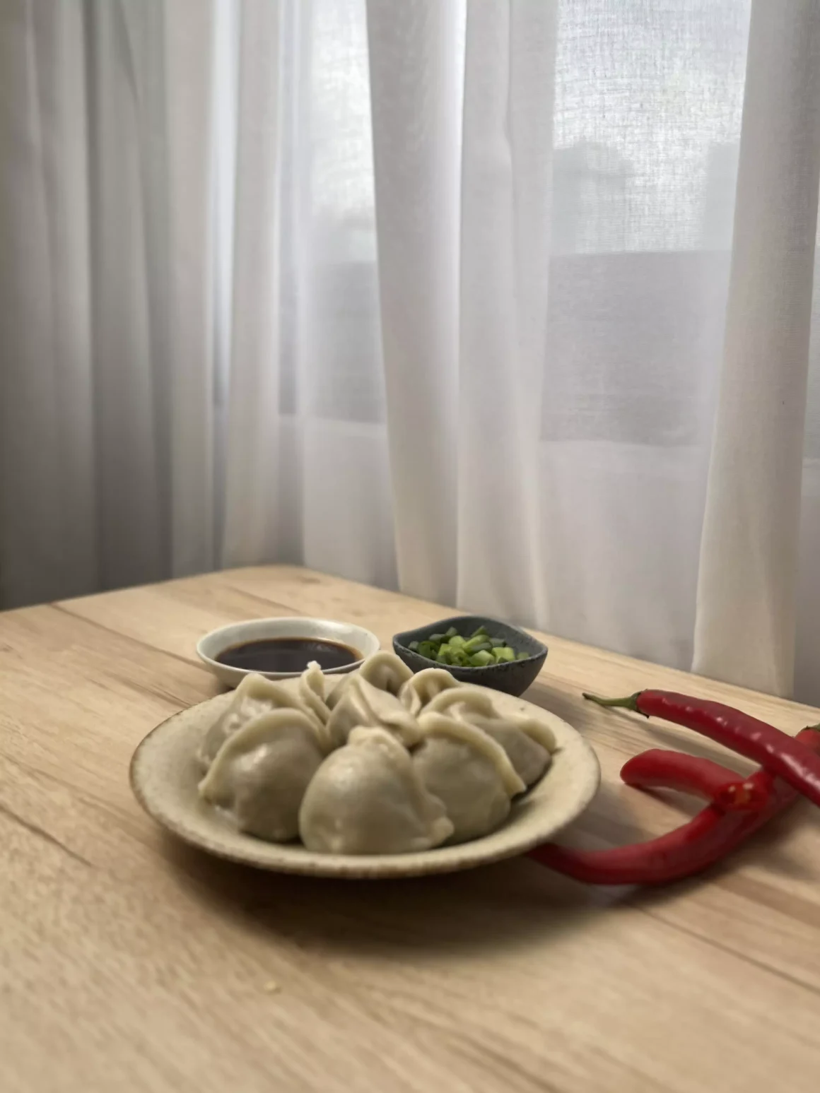 《猿山鹿》水餃好吃嗎?水餃界的掃地僧,一個臥底KTV多年的秘密武器開箱 6 一盤剛煮好的猿山鹿水餃擺放在木紋桌上,旁邊搭配著醬油、蔥花和新鮮的紅辣椒,窗外光線柔和。